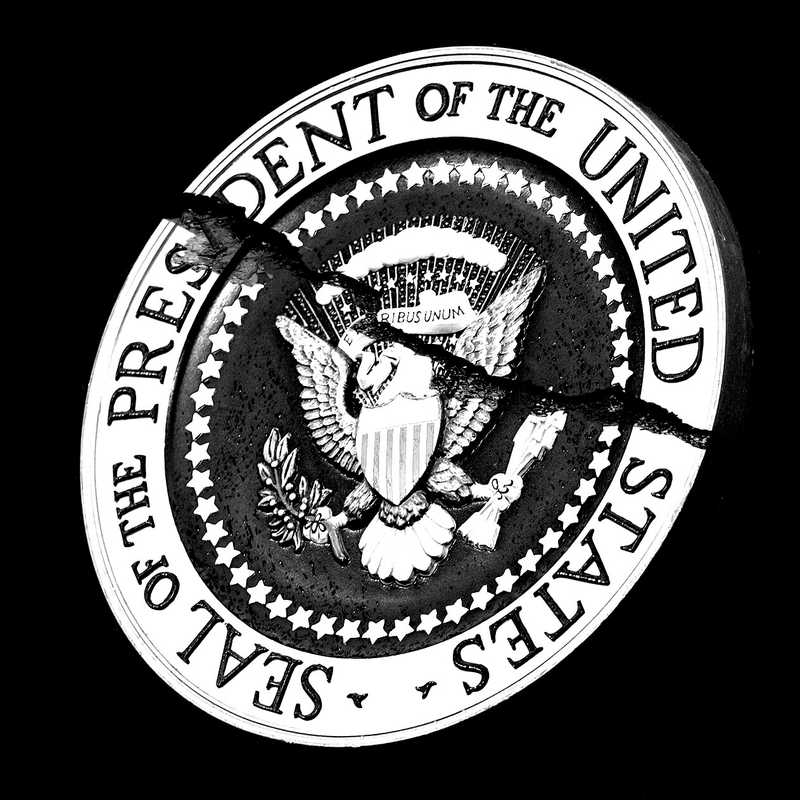 A photo illustration of the presidential seal, which features an eagle holding an olive branch in its left talon and a bundle of arrows in its right. In the center of the crest is a shield and above the eagle are the words 'E pluribus unum.' The crest is surrounded by stars. The image is in black and white with a large crack splitting the seal diagonally in half.